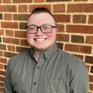 A masculine person with brown hair wearing a green shirt and black glasses against a brick wall