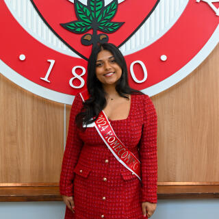A smiling woman in a red suit dress stands before the seal of Ohio State University