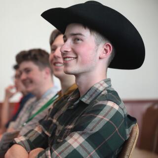 A smiling person in profile wearing a plaid shirt and a cowboy hat