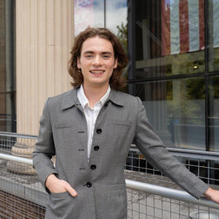 A smiling person in a white shirt and grey jacket