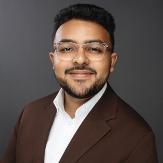 A smiling man in a white shirt, brown jacket and glasses