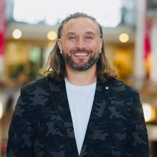 A smiling man with long hair in a white shirt and black camouflage suit jacket