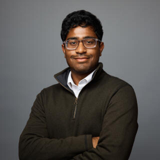 A smiling man in a white shirt, green sweater and glasses with his arms crossed