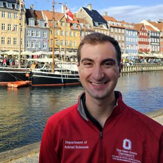 A smiling man in a red Ohio State jacket stands before a canal and row houses in Copenhagen