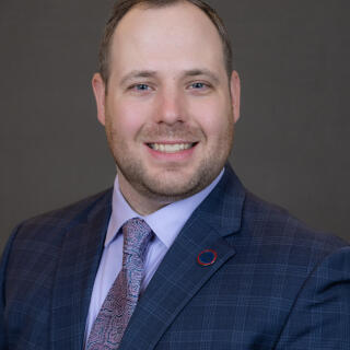 A smiling person in a blue shirt, jacket and tie with a round lapel pin