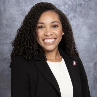 A smiling woman in a white shirt and black jacket with a Block O lapel pin