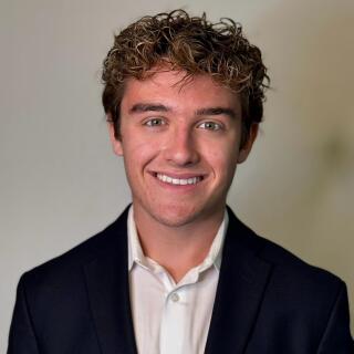 A smiling man in a white shirt and black jacket with a Block O lapel pin