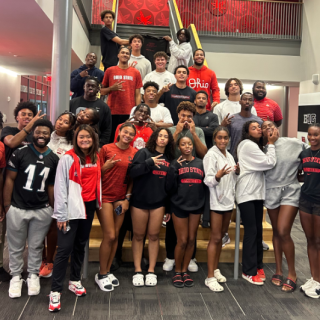 A group of smiling people stand together before a set of indoor stairs