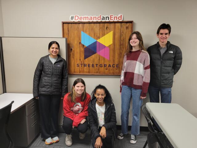 Five college student volunteers posing in front of wood sign with Street Grace logo