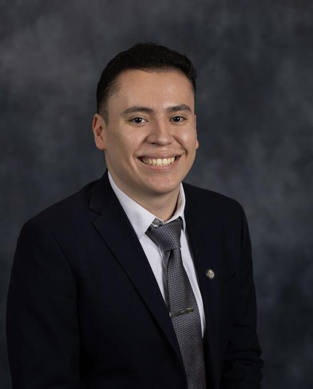 A headshot image of Alex Ochoa. He has brown hair and brown eyes, and wears a black suit.