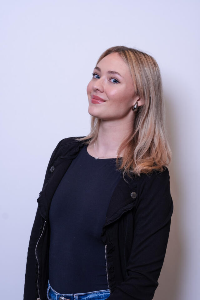 An image of Ava Wohner leaning against a white wall. She has blonde hair and blue eyes, wearing a black top.