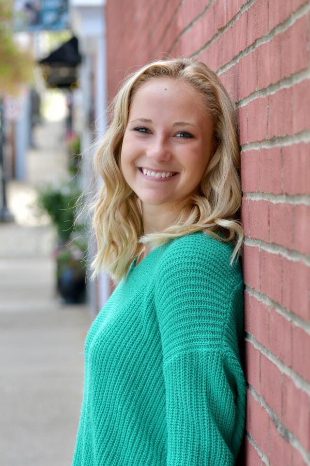 An image of Brooke Tacsar leaning against a brick wall. She has blonde hair and is wearing a teal sweater.