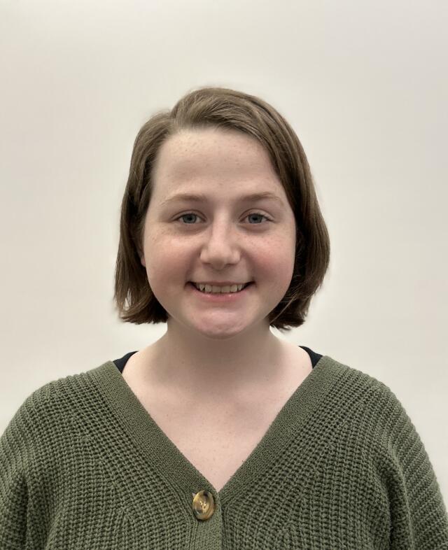 Headshot image of Callie Thayer in front of a white wall. She has brown hair and brown eyes and is wearing a green sweater.