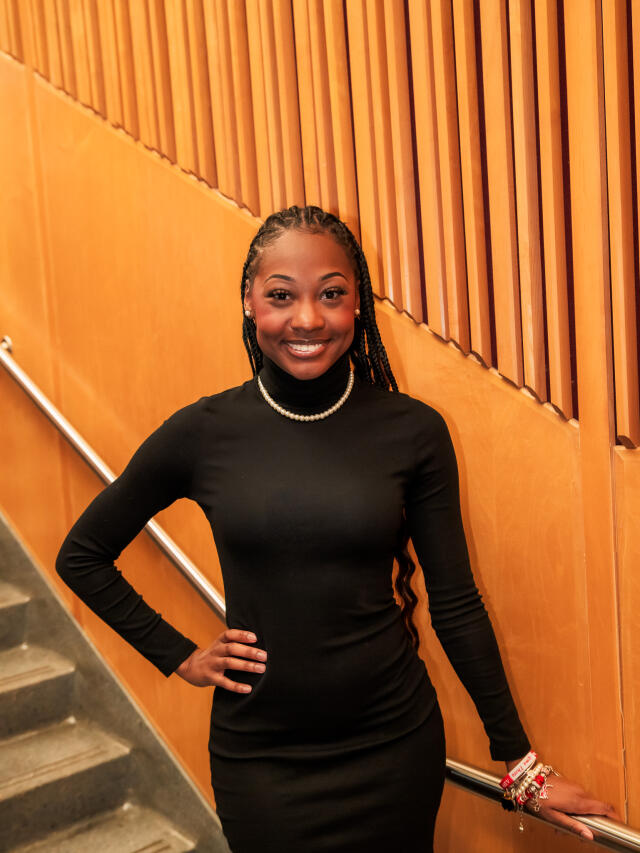 An image of Camryn Little posing in front of a stair case. She has brown skin, long braids and is wearing a black turtleneck dress.