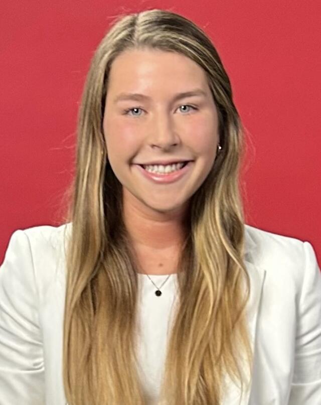 A headshot image of Clara Planner in front of a red backdrop. She has blue eyes and long blonde hair, and wears a white blazer.