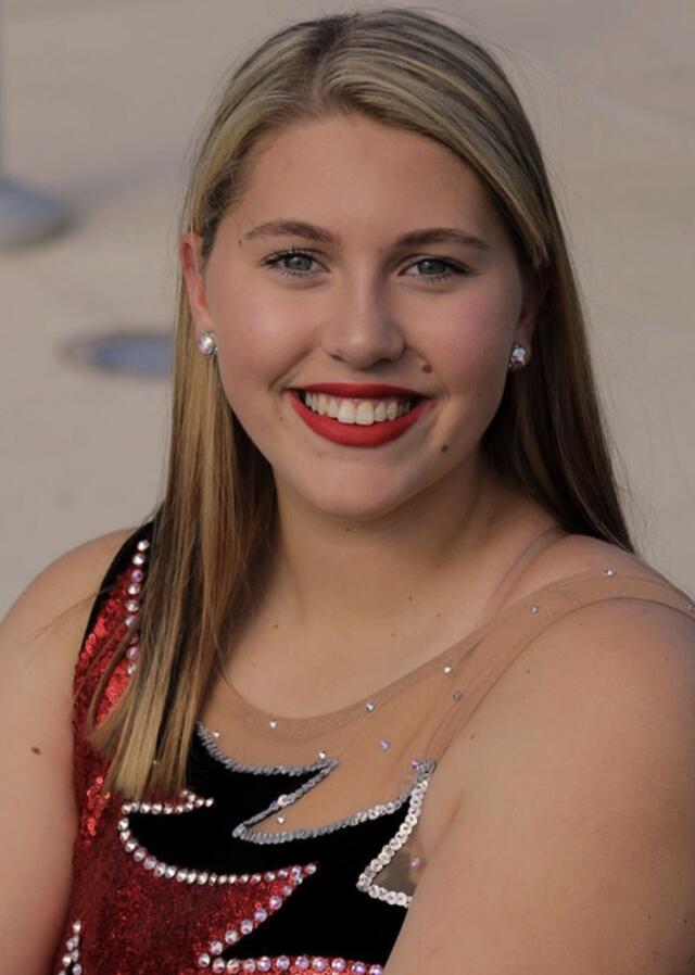 A headshot image of Erica Kauffman outdoors. She has medium-length blonde hair and blue eyes, and wears a dance uniform.