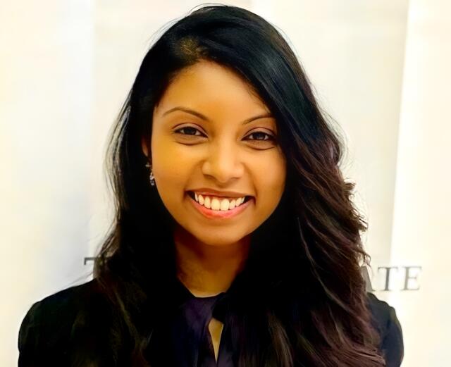 An headshot image of Iresha Jayasinghe in front of a white wall, she has dark brown hair and is wearing a black top.