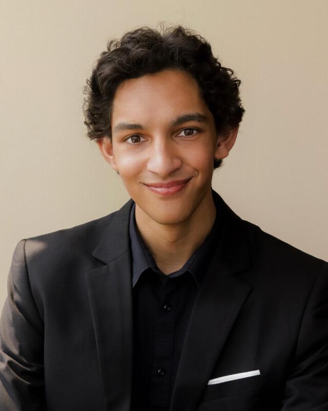 A headshot image of Joshua Ghosh-Groen. He has styled dark brown hair and wears a black blazer with a black button down shirt.