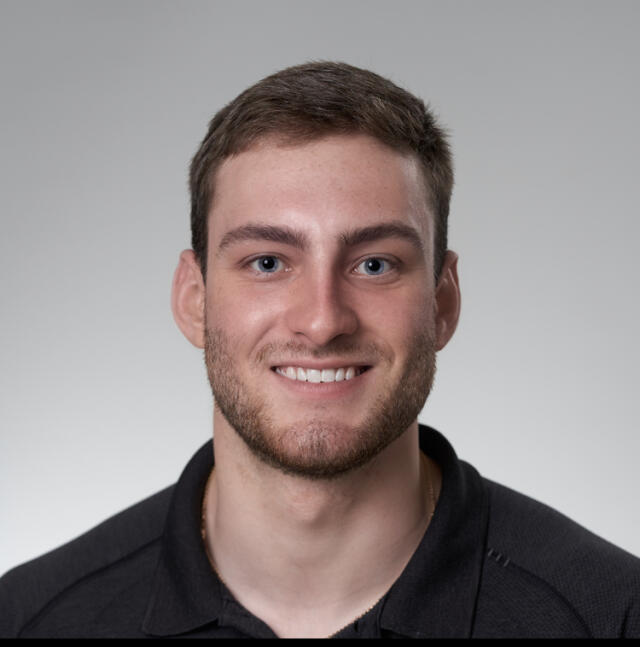 A headshot image of Mason Myers in front of a gray background. He has short brown hair and is smiling. He wears a black polo shirt.