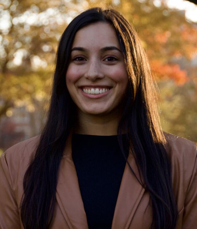 An image of Monica Pannett outdoors. She has long brown hair and is smiling. She wears a brown blazer with a black top underneath.