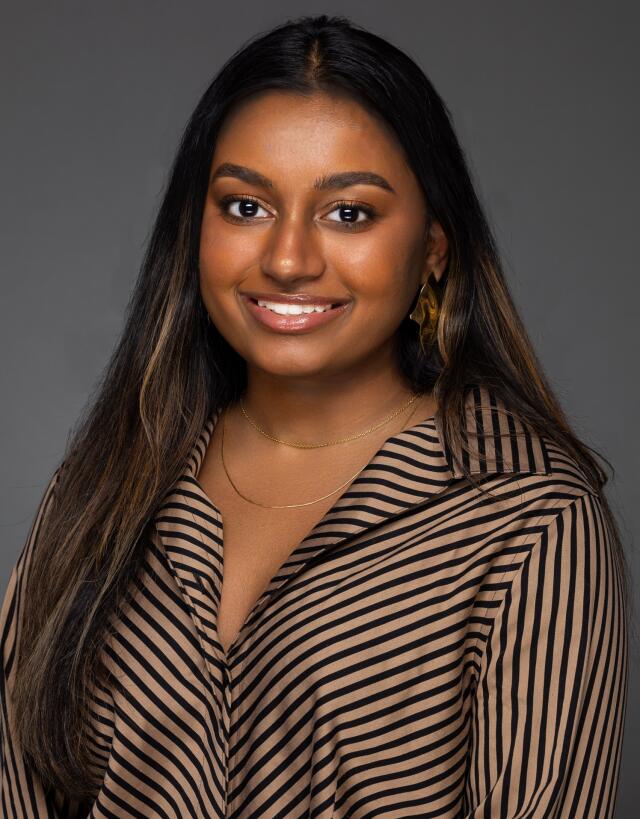 A headshot image of Prateeksha Prabhakar. She has long, brown hair with blonde highlights and brown skin. She wears a striped shirt.