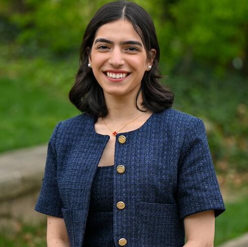 An image of Sara Alsammerai outdoors. She has short, dark brown hair and is smiling. She wears a navy blue tweed blazer with a matching skirt.