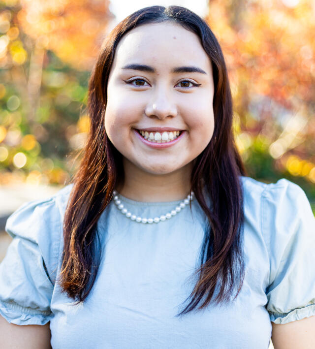 An image of Regina Loayza outdoors. She has brown hair and brown eyes, and wears a striped shirt.