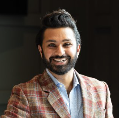 A smiling man in a blue shirt and a red and brown plain jacket