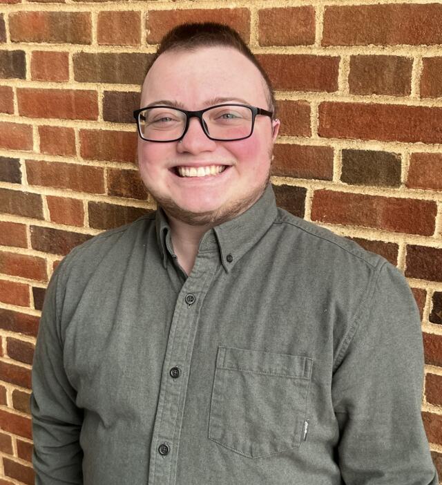 A masculine person with brown hair wearing a green shirt and black glasses against a brick wall