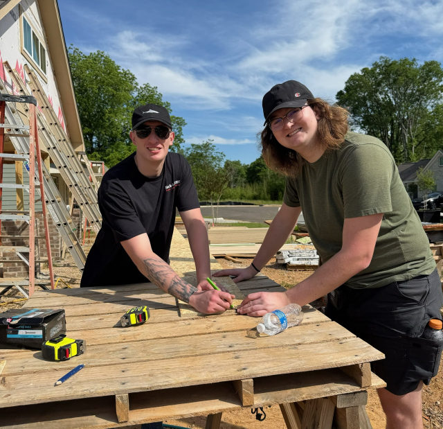 Two students measure a piece of wood to cut on a platform supported by two sawhorses