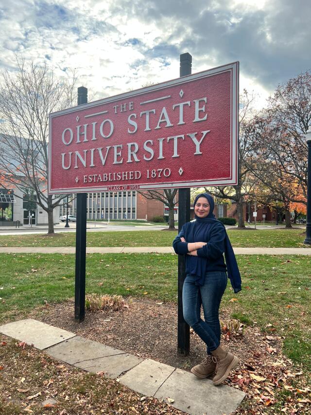A smiling woman standing with her legs crossed next to a sign reading The Ohio State University
