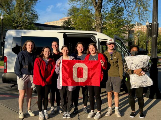 8 college students standing outside in front of white van and holding an OSU flag