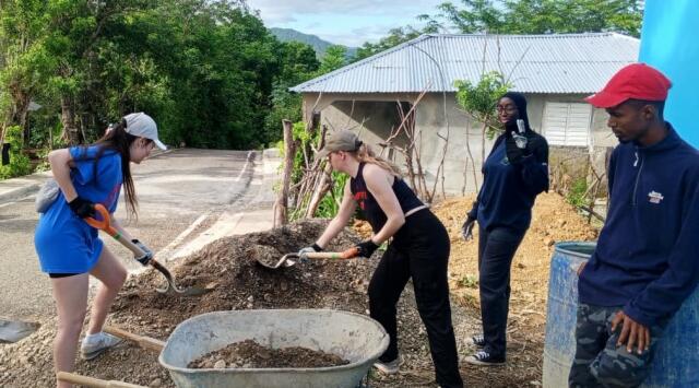 Two students shovel construction material into a wheelbarrow while two others look on.