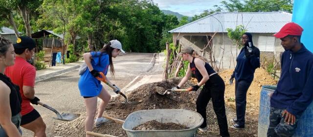 Three volunteers work to shovel dirt into a wheelbarrow while 3 other volunteers wait to assist.