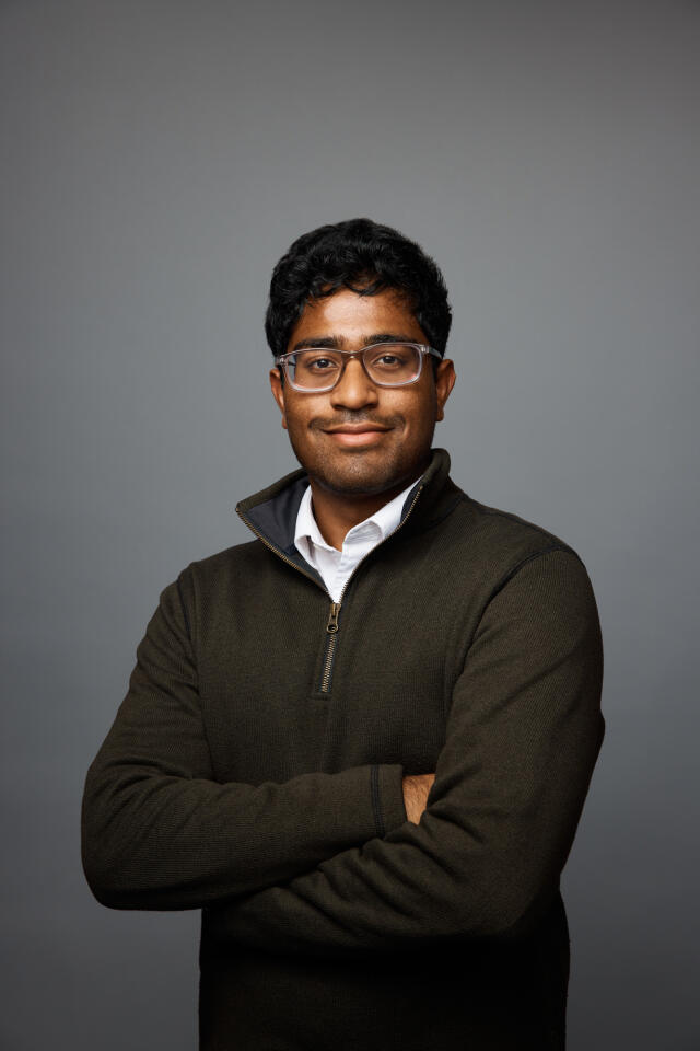 A smiling man in a white shirt, green sweater and glasses with his arms crossed