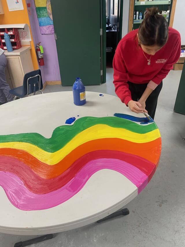 Volunteer wearing red shirt painting a rainbow pattern on the surface of a round table