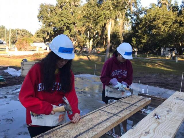 Two college student volunteers with protective glasses and hard hats hammering planks of wood
