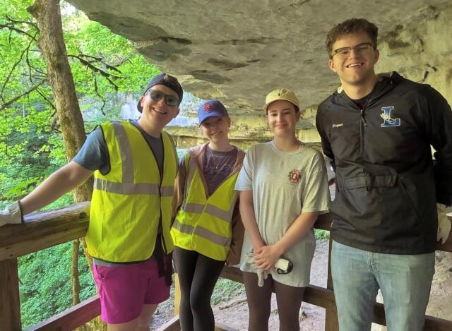 Four participants pose on a raised platform with a cave and rock formation behind them