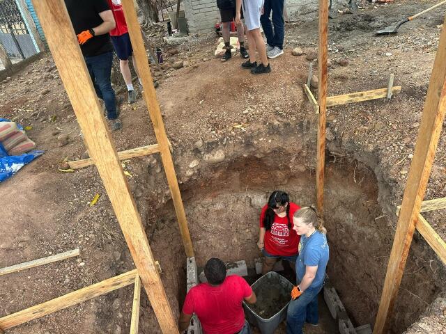 Three student volunteers are in a hole deeper than they are tall, working to remove dirt