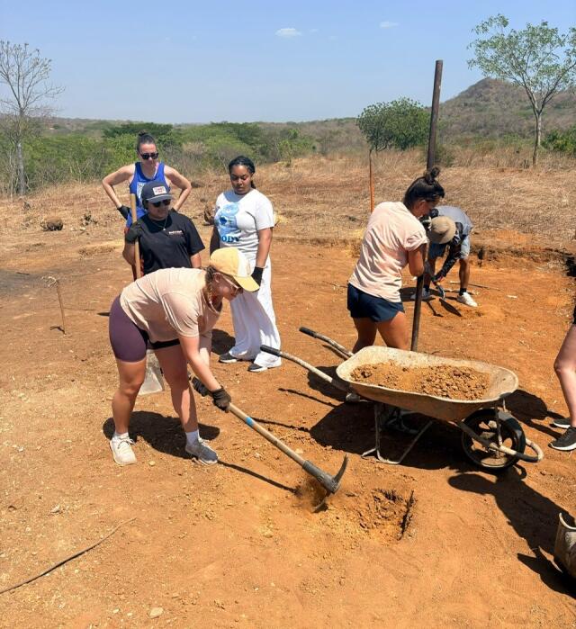 5 students work to clear a worksite using pick axes and shovels, moving dirt into a wheelbarrow