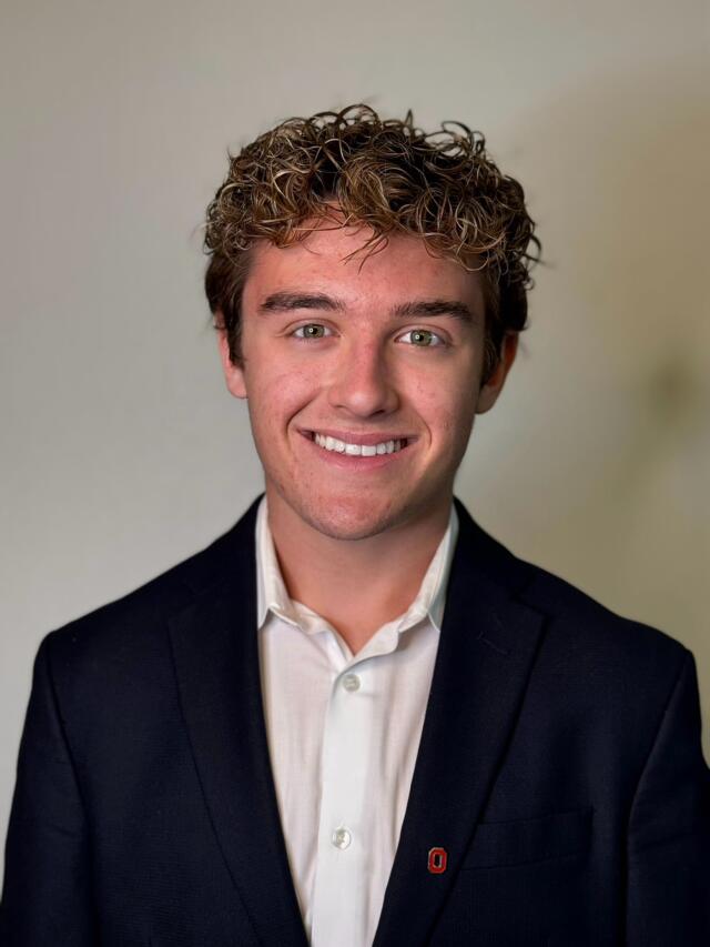 A smiling man in a white shirt and black jacket with a Block O lapel pin