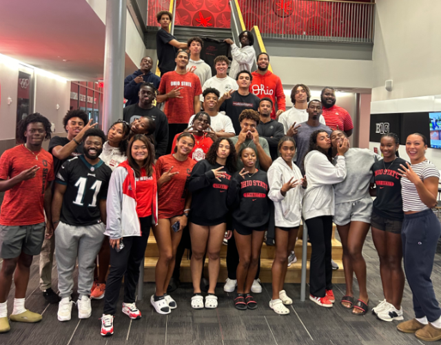 A group of smiling people stand together before a set of indoor stairs